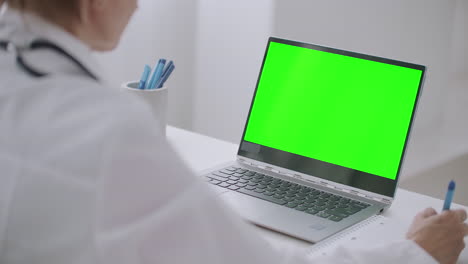 Eine-Virologin-Schaut-Sich-Ein-Online-Webinar-An,-Schaut-Sich-Das-Grüne-Display-Eines-Laptops-Für-Die-Chroma-Key-Technologie-An,-Schreibt-Notizen-Auf-Papier-Und-Nickt